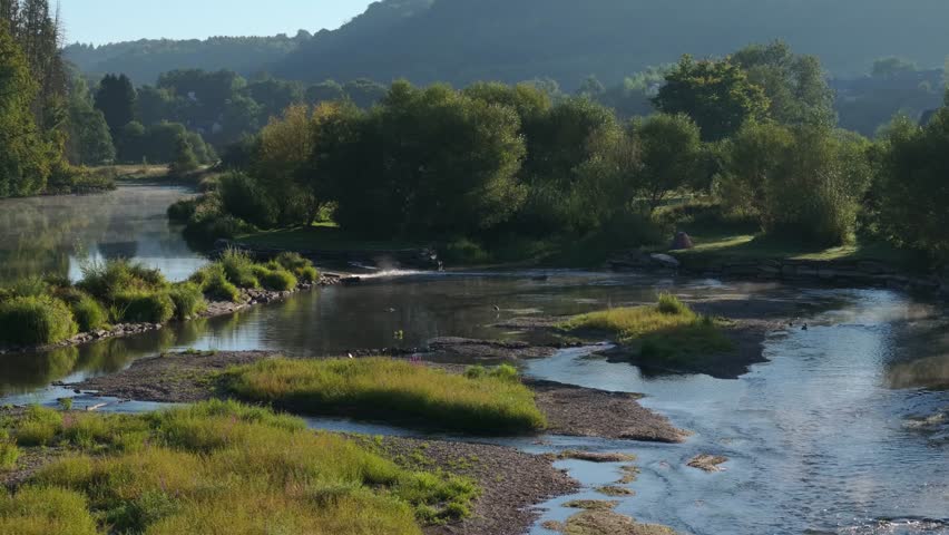 Golden hour sunlight shines over river with rocks, trees, and buildings near Moulin de Cugnon