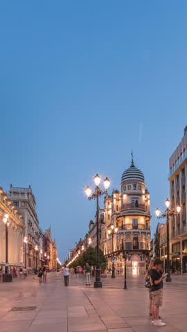 Avenida de la Constitucion day to night timelapse in Seville, Spain. Historic architecture with modern streetlights, pedestrians and urban atmosphere. Iconic Adriatica building glowing after sunset.