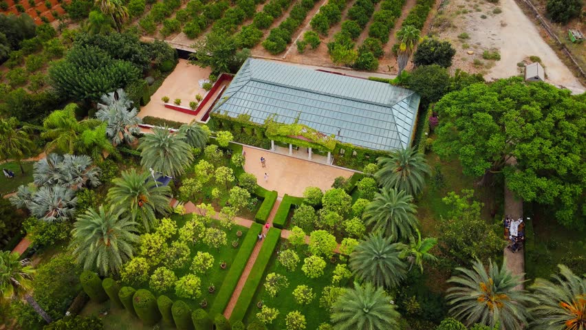 Drone video shows a lush public garden in Spain, with structured greenery, a pool, and architectural elements surrounded by orchards and scenic landscapes.
