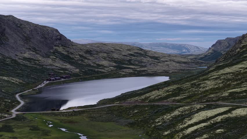 An aerial footage of a scenic still lake reflects cloudy skies, cradled by soft green ridges in Rondane National Park, Norway