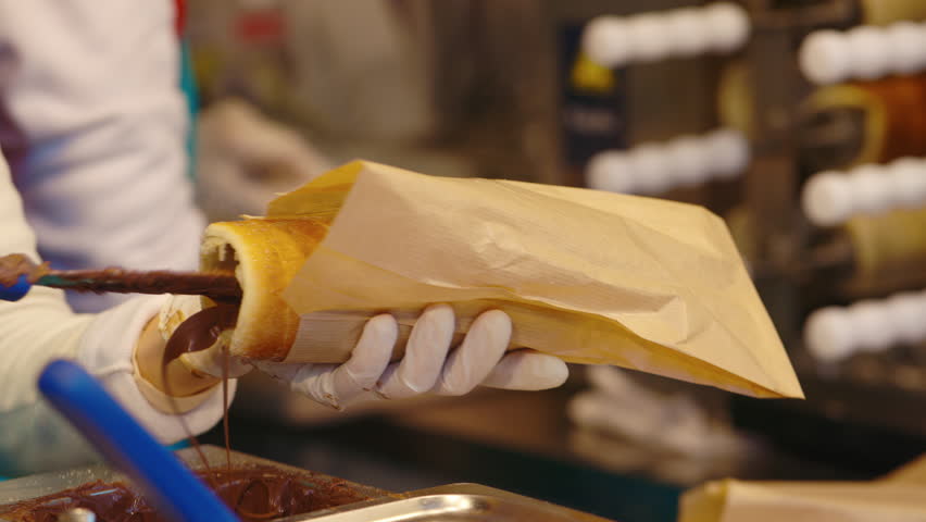 Closeup view of woman in gloves fills traditional trdelnik with chocolate paste in winter fair stall. Baker prepares tasty Austrian dessert at Christmas holiday outdoors. Fast food