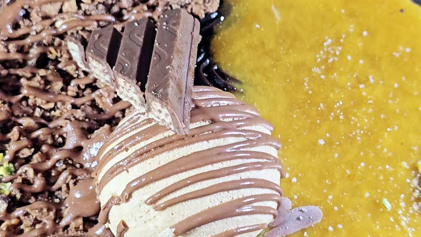 Ice cream cake, on a sponge cake layer, topped with chocolate pieces and cream and sauce, chunks of chocolates with pistachio pieces, half mango and half chocolates, summertime and holidays concept