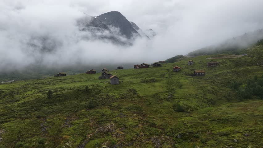 An aerial footage of the misty mountains loom over scattered wooden cabins in a lush green valley in Skjerdingsdalssaetre, Videseter,Norway