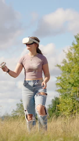 Vertical shot of woman walking while wearing virtual VR headset in green grassy field