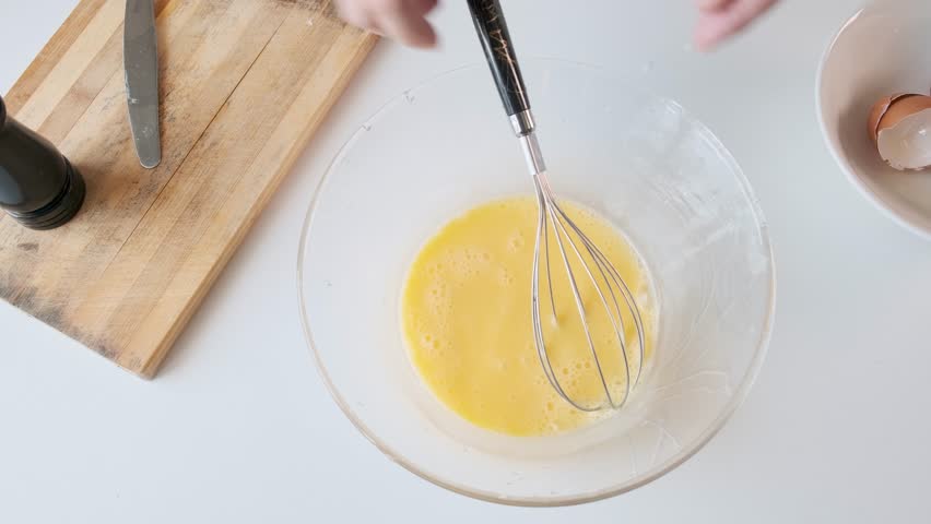 Man seasoning and whisking beaten eggs in glass bowl with pepper grinder in kitchen with milk during breakfast preparation. Cooking omelette or scrambled eggs. Healthy food. Top view