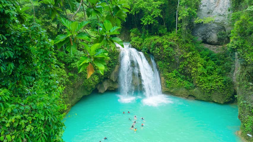People swimming and enjoying the clear turquoise water of a multi-tiered waterfall, surrounded by dense tropical jungle foliage, Kawasan Falls, Cebu, Philippines.