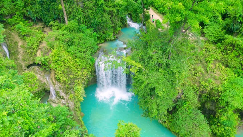 Aerial view of a multi-tiered waterfall cascading into vivid turquoise pools surrounded by dense green tropical forest. Kawasan Falls, Cebu, Philippines.