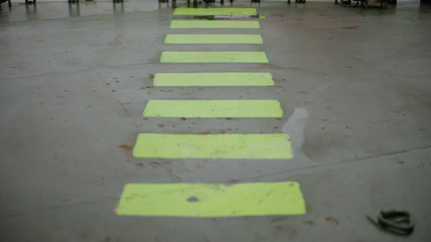 Fluorescent safety walkway markings on a polished concrete floor in an industrial facility, forming a bright guided path with reflective wet patches and strong perspective lines.