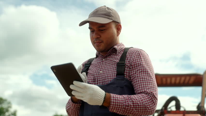 Confident farmer with tractor in crop field, using tablet and smart farming technology for crop monitoring, precision agriculture, and modern farm management.
