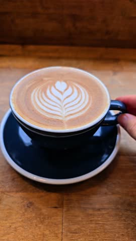Vertical video capturing a close-up of a hand holding a dark blue coffee cup with a saucer, showcasing elegant leaf-shaped latte art made with steamed milk. The cup rests on a wooden table 