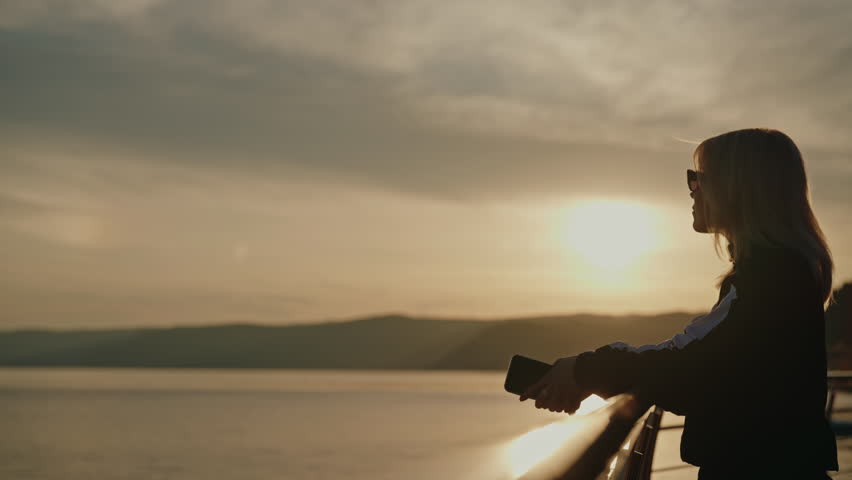 Relaxed blonde woman in sunglasses holding smartphone, leaning on railing of pier, enjoying breathtaking view of tranquil lake and rolling hills during picturesque sunset. Concept of vacations, travel