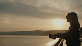 Relaxed blonde woman in sunglasses holding smartphone, leaning on railing of pier, enjoying breathtaking view of tranquil lake and rolling hills during picturesque sunset. Concept of vacations, travel - Powered by Shutterstock - Get 15% off with code: PIKWIZARD15