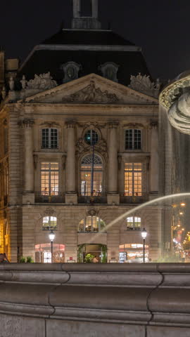 View of Place de la Bourse and the fountain at night timelapse in Bordeaux, France. Illuminated historic buildings border the large square. Glowing windows in a restaurant