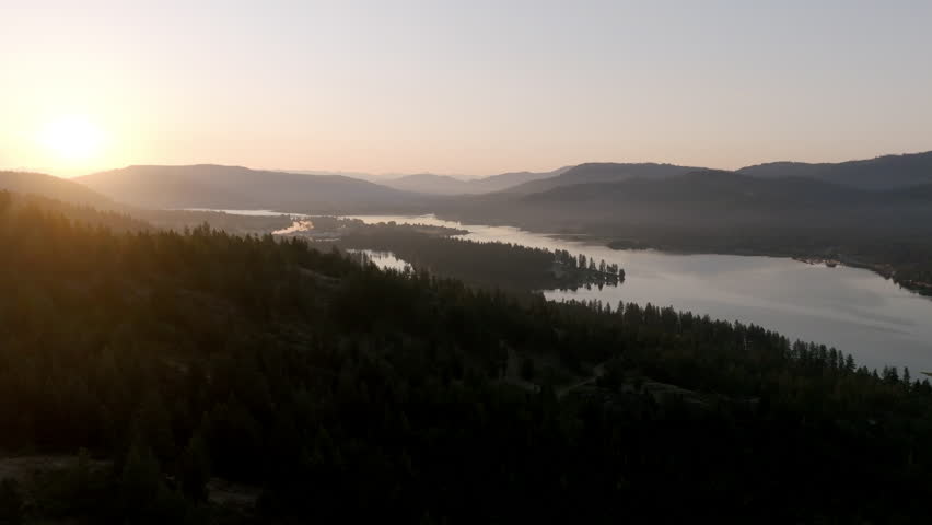 The sun crests over distant mountains in Priest River, Idaho, illuminating reflective lakes and dark forests, a peaceful wilderness waking to golden morning light.