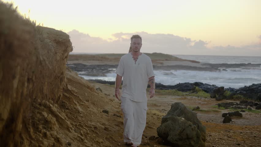 A man in casual white clothing walks toward the camera along a rugged coastal path, with waves breaking against rocks under soft golden sunrise light.
