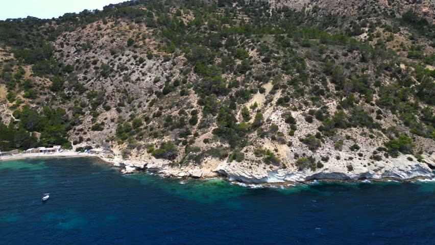 Aerial view of boats mooring near a rocky coast with lush vegetation in Ibiza, Spain. Fantastic aerial view flight rotation pan to right drone