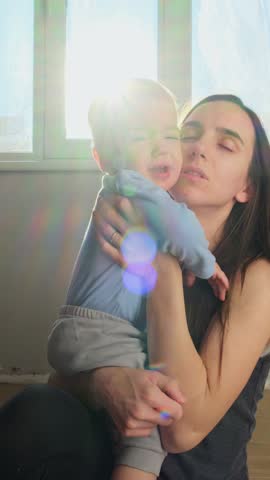 Mother calms the child in teeth pain and hugs her crying baby toddler early in morning. Warm family moment of care, empathy, and affection , sunshine on background