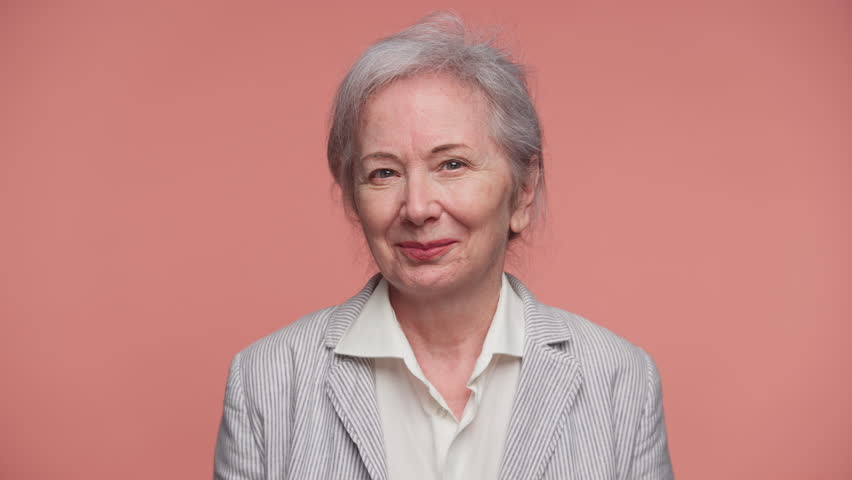 Confident elderly woman winks with a slight smile, expressing charm and humor while facing the camera against a pink background.