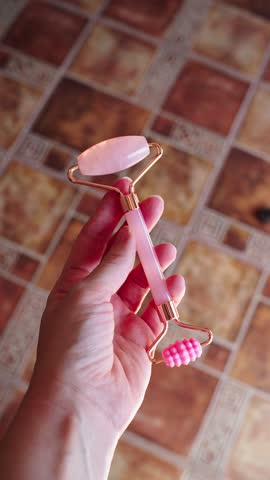 A person is holding a pink quartz facial roller in one hand. Sunlight filters through, illuminating the roller in a cozy indoor setting.