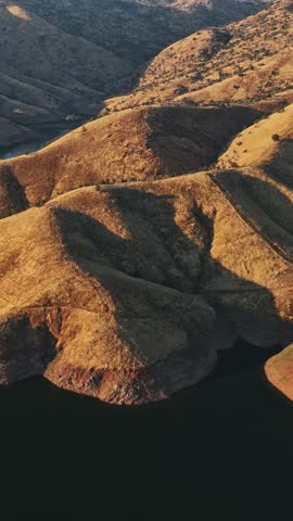 The aerial perspective showcases the distinct terrain of California, highlighting the undulating hills that border a serene water body under clear skies.
