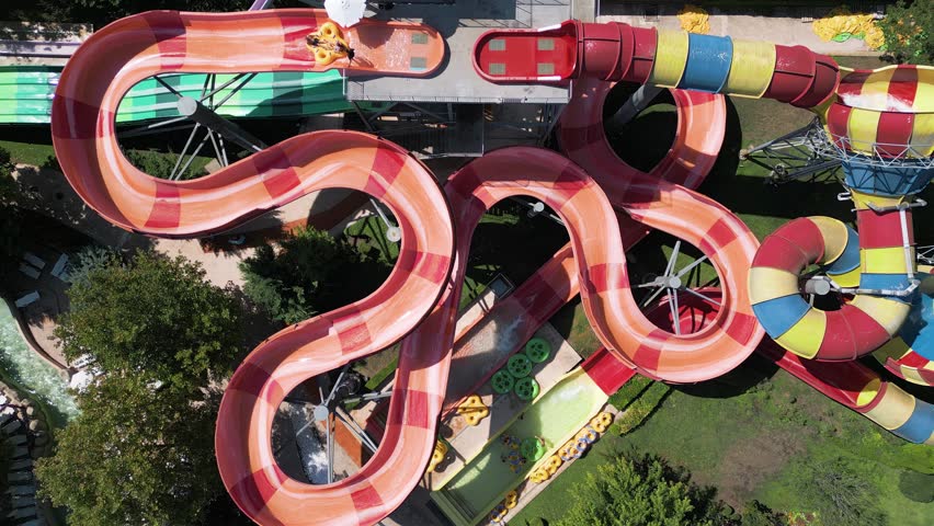 Aerial view of colorful twisting water slides with people riding inflatable tubes at a water park. Bright summer day filled with thrill, fun, and excitement