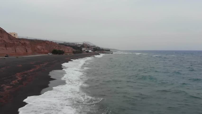 Aerial footage capturing the unique black sand beach and rugged volcanic coastline of Santorini island, Greece.
