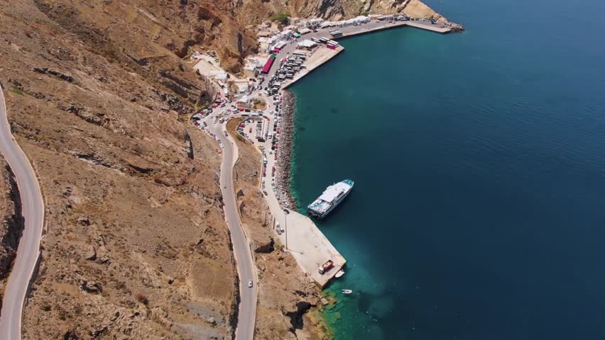 Drone footage captures the stunning aerial perspective of a bustling port on Santorini Island, Greece, featuring a winding coastal road, vibrant blue Aegean Sea, and traditional white-washed buildings