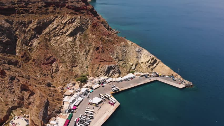 Aerial footage showcasing the bustling Athinios Port in Santorini, Greece, with ferries and boats docked against the dramatic volcanic cliffs and the serene Aegean Sea.