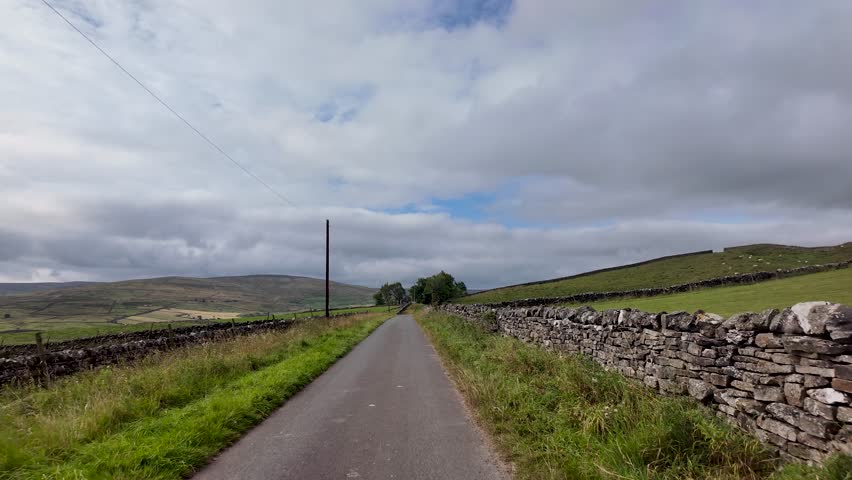 A narrow country road winding through vast green landscape with stone walls and rolling hills.