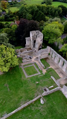 Mid-angle aerial drone pans left over Bishop’s Waltham Palace ruins nestled among autumn trees, capturing historic stone walls and warm golden sunset light for a scenic village landscape.