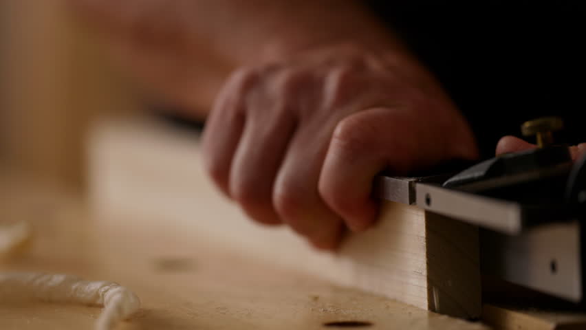 Craftsman focuses on shaping a wooden piece using a hand plane, demonstrating skilled woodworking techniques in a bright, organized workshop setup.