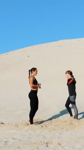 Two athletic, young women in black fitness suits are engaged in a pair, work out kicks, train, perform squats and kick-ups, on deserted beach, in summer, under a hot sun.
