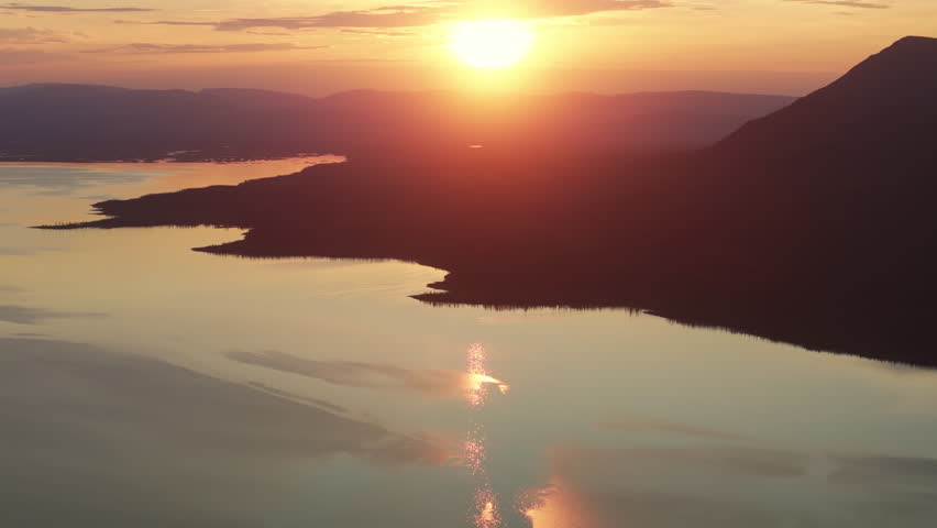 Aerial view of the sun glowing above Lake Lama and the rugged shoreline. A long reflection stream leads toward layered ridges and endless taiga across the Putorana Plateau in Krasnoyarsk Krai, Russia