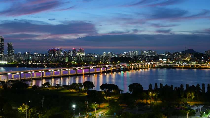 Zoom out time lapse of the famous Banpo Bridge Rainbow Fountain show at sunset, revealing the Han River Park and fast moving car traffic light trails and Seoul panoramic cityscape - aerial
