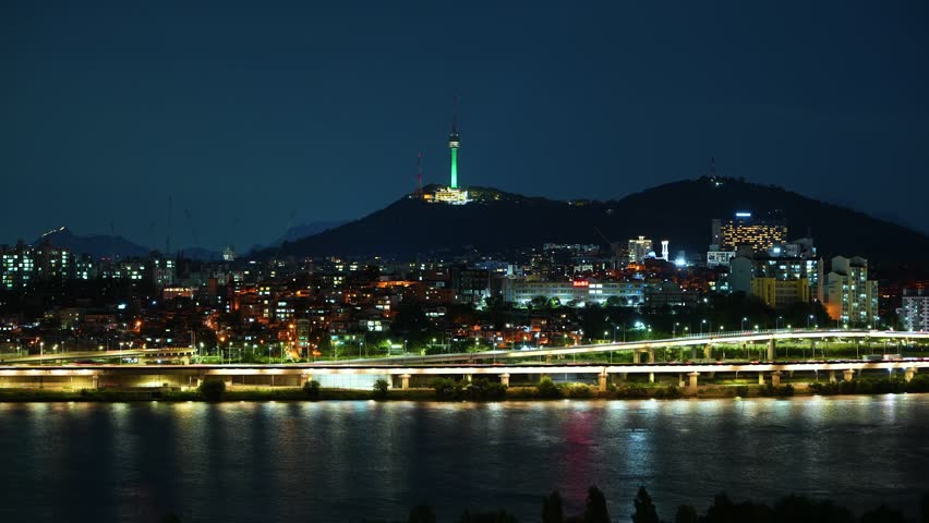 Zoom in time lapse of the iconic Namsan Tower and the vibrant Seoul cityscape at night, with glowing city lights and fast moving traffic on expressway next to Han river creating light trails on