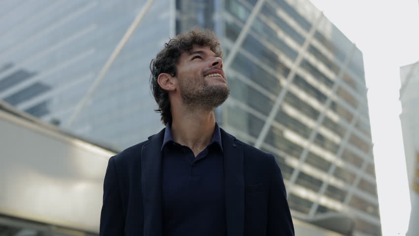 Portrait of Hispanic man office employee smiles and looks upward in urban landscape. Ambitious Latin man entrepreneur dreams about success on downtown street. Big career plans