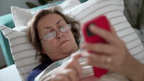 Elderly woman lying in bed using a red smartphone, illustrating the digital engagement and modern lifestyle of seniors in a relaxed home atmosphere - Powered by Shutterstock - Get 15% off with code: PIKWIZARD15