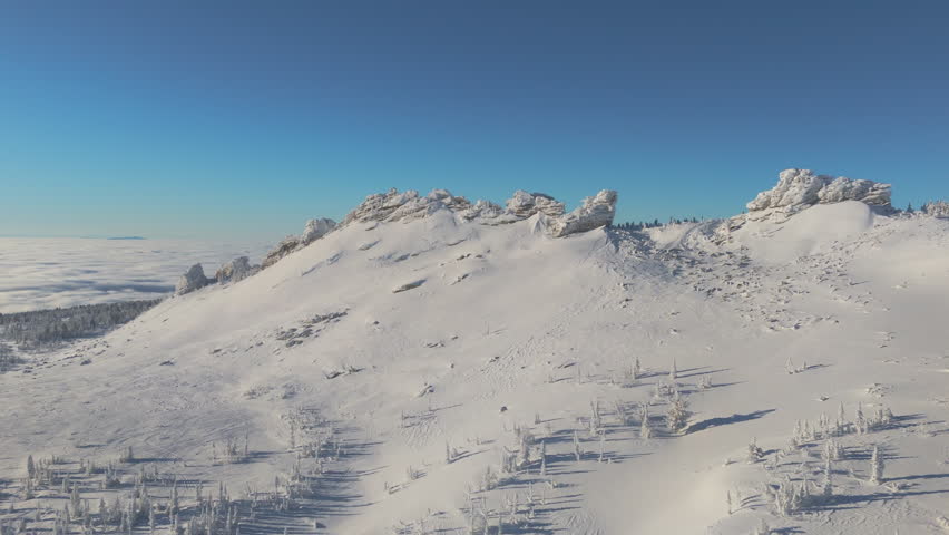 Drone glides along a wind-carved rock ridge above the taiga of Sheregesh, crystalline snow covering the plateau under a cloudless sky. Massive blocks and cornice detail add drama and geologic scale