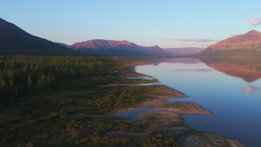 Aerial view of Lake Lama coastline at blue hour with pink mountain reflection across transparent water. Gentle river mouth, taiga margin and clear sky deliver rare Arctic travel atmosphere