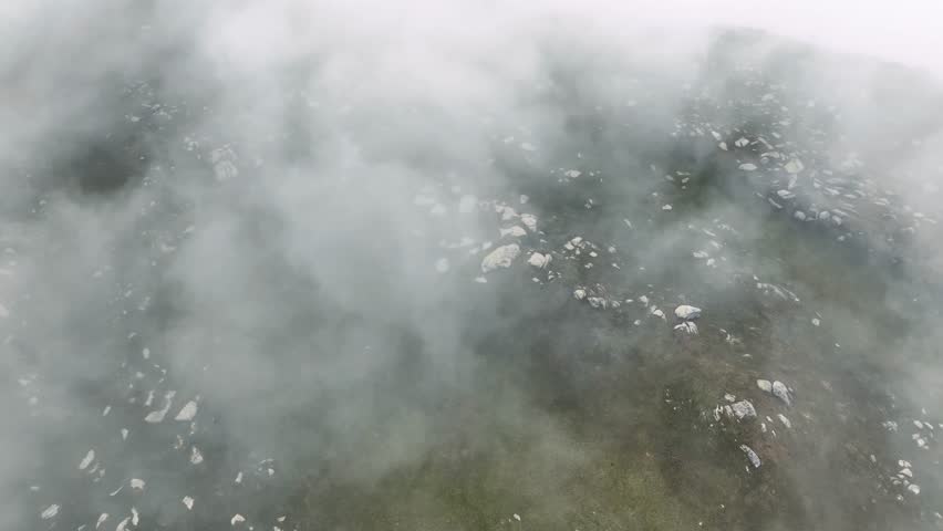 A lone hiker in an orange jacket stands on a rocky mountain summit surrounded by dense fog, symbolizing adventure and solitude.
