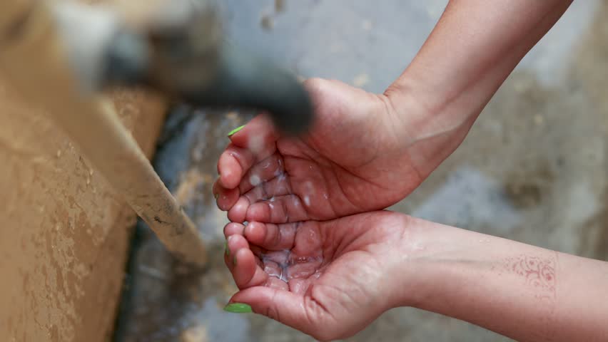A top-view shot of water dripping onto a woman’s cupped palms from a tap, symbolizing water scarcity in rural areas of developing countries. A powerful reminder of limited resources.