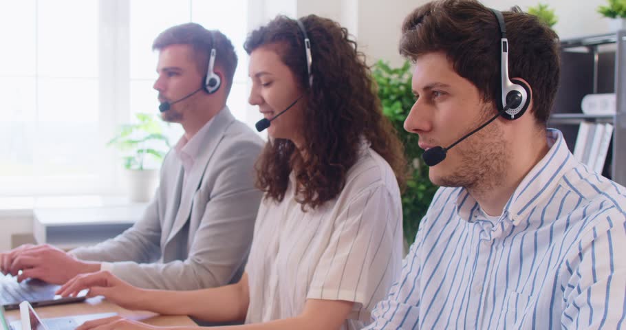Three young customer support services employees wearing headsets consulting clients seated at desk in modern office, providing help, resolving issues through phone or online chat support