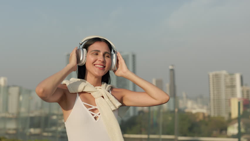Portrait of Brazilian woman with headphones listens to music and smiles on embankment. Joyful young lady dances expressing good mood against distant downtown skyscrapers in city