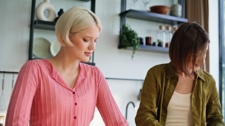 Smiling roommates standing kitchen arranging vegetables in bright early daylight closeup. Excited couple sharing peaceful breakfast time indoors. Energetic girl cutting greens friend making granola