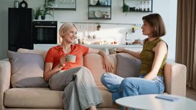 Lgbt couple talking sofa enjoying cozy atmosphere. Smiling two women drinking beverages chatting calmly in modern apartment. Pair lesbian girls have friendly conversation embracing together at house - Powered by Shutterstock - Get 15% off with code: PIKWIZARD15