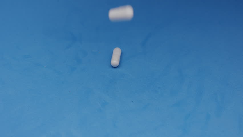 White capsules or pills being poured from a plastic bottle into a hand against a blue background. Concept of medicine, healthcare, supplements, and pharmaceutical treatment.