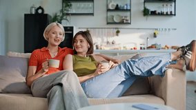 Happy lgbt couple drinking coffee together on sofa cozy living room. Relaxed women bonding at peaceful home morning. Joyful girlfriends talking sitting couch enjoying comfortable domestic atmosphere - Powered by Shutterstock - Get 15% off with code: PIKWIZARD15