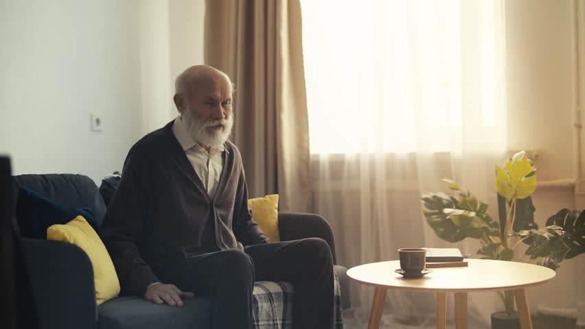 Senior man feeling sudden knee pain while standing up from couch, rheumatism