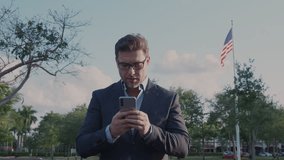 Businessman in suit reads breaking news on smartphone and reacts with excitement. Male executive stand outdoors with American flag behind, symbolizing politics, economy, Wall Street and global finance - Powered by Shutterstock - Get 15% off with code: PIKWIZARD15
