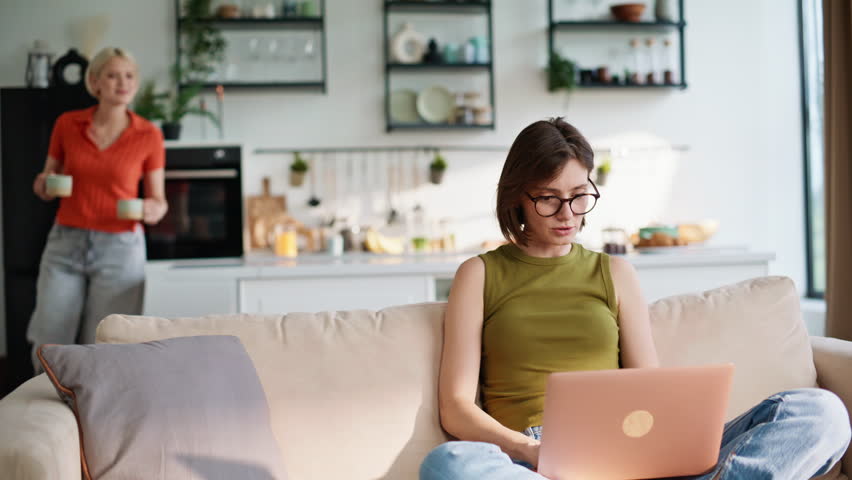 Focused woman working laptop cute girlfriend bringing cup of coffee embracing closeup. Lgbt couple spending time at home together. Brunette lady watching computer screen talking to positive bestie